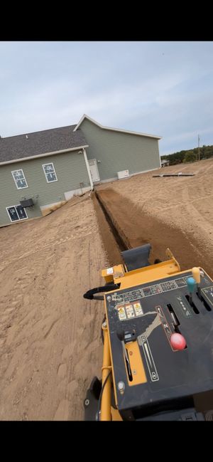 Construction trench beside a house, with a yellow machine and black tool panel in the foreground.