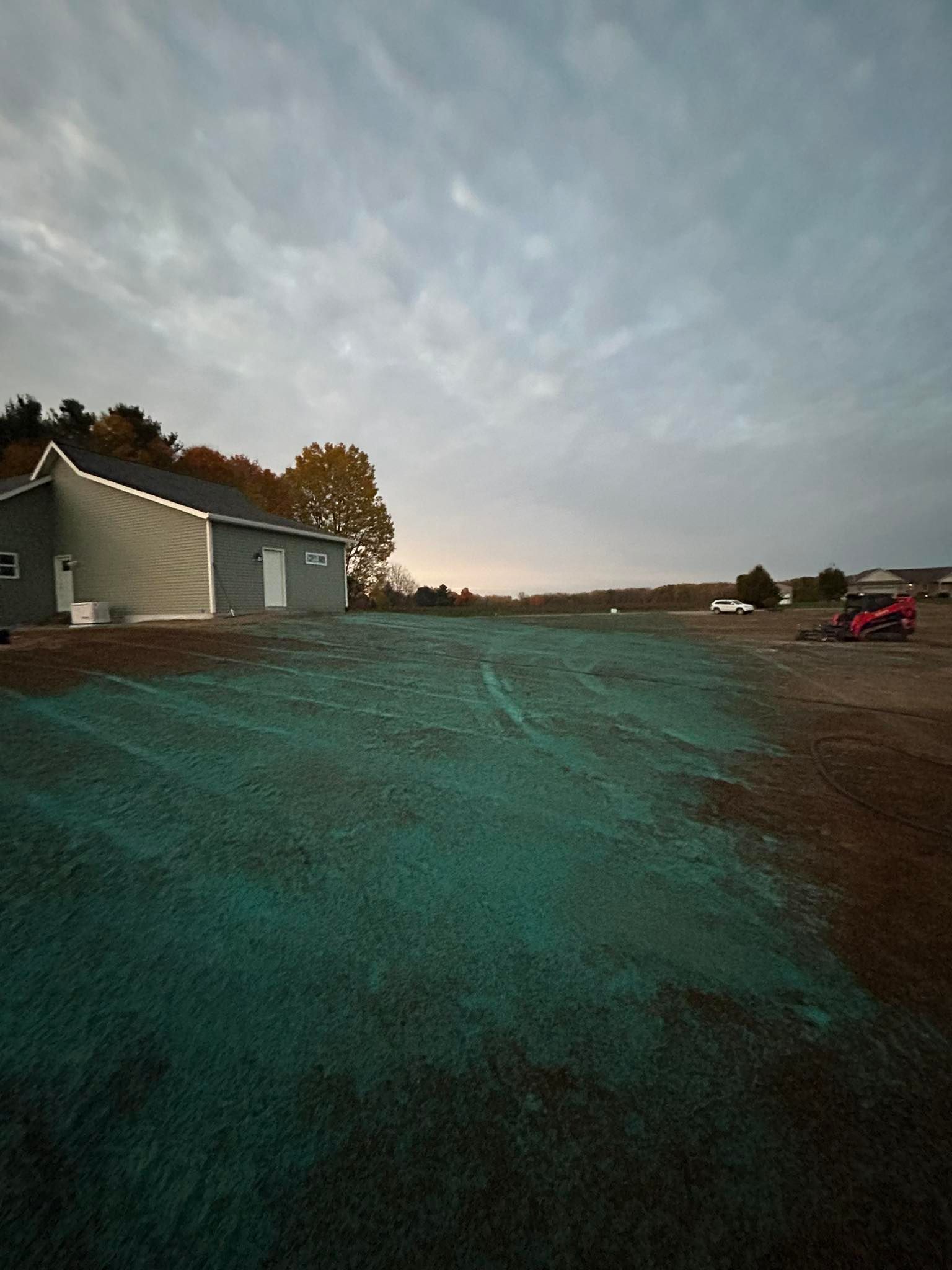 Wide rural yard at dusk beside a gray house, with a pink vehicle in the distance under a cloudy sky