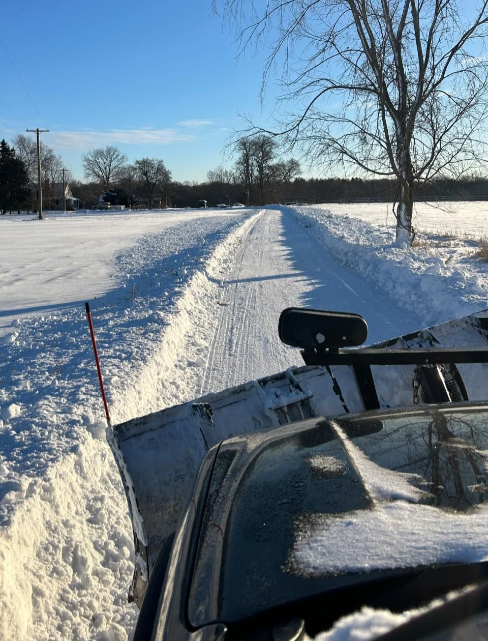 Snow-covered path beside a frozen field, viewed from a vehicle plowing through deep snow.