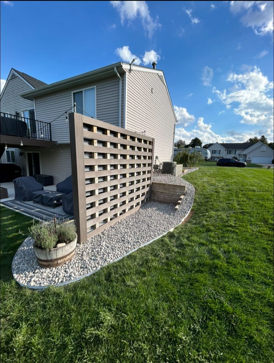 Backyard patio with beige privacy lattice, potted plants, rocks, and green lawn beside a house.