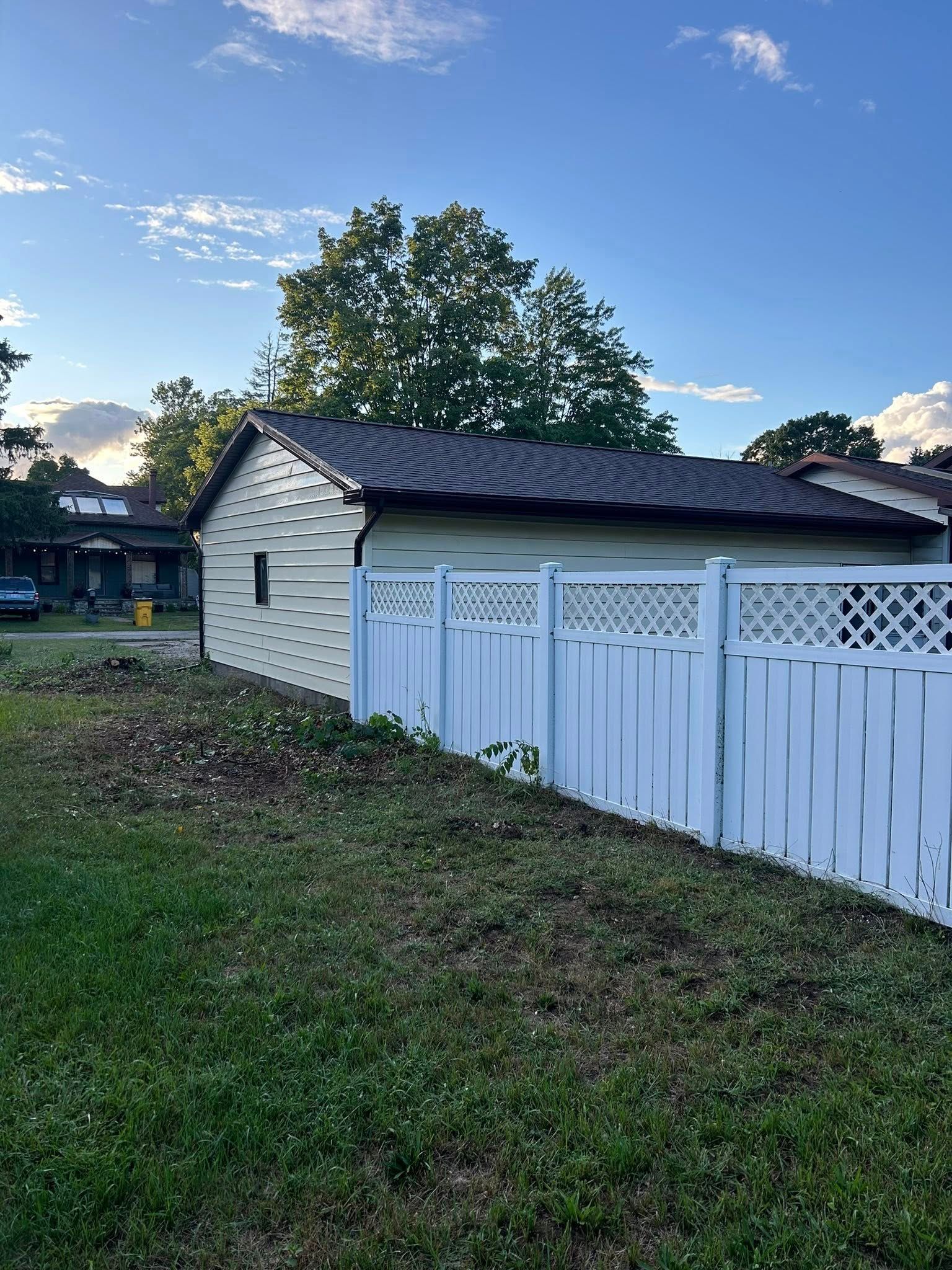Small white shed beside a white fence in a grassy yard at dusk, with trees and houses in the background.