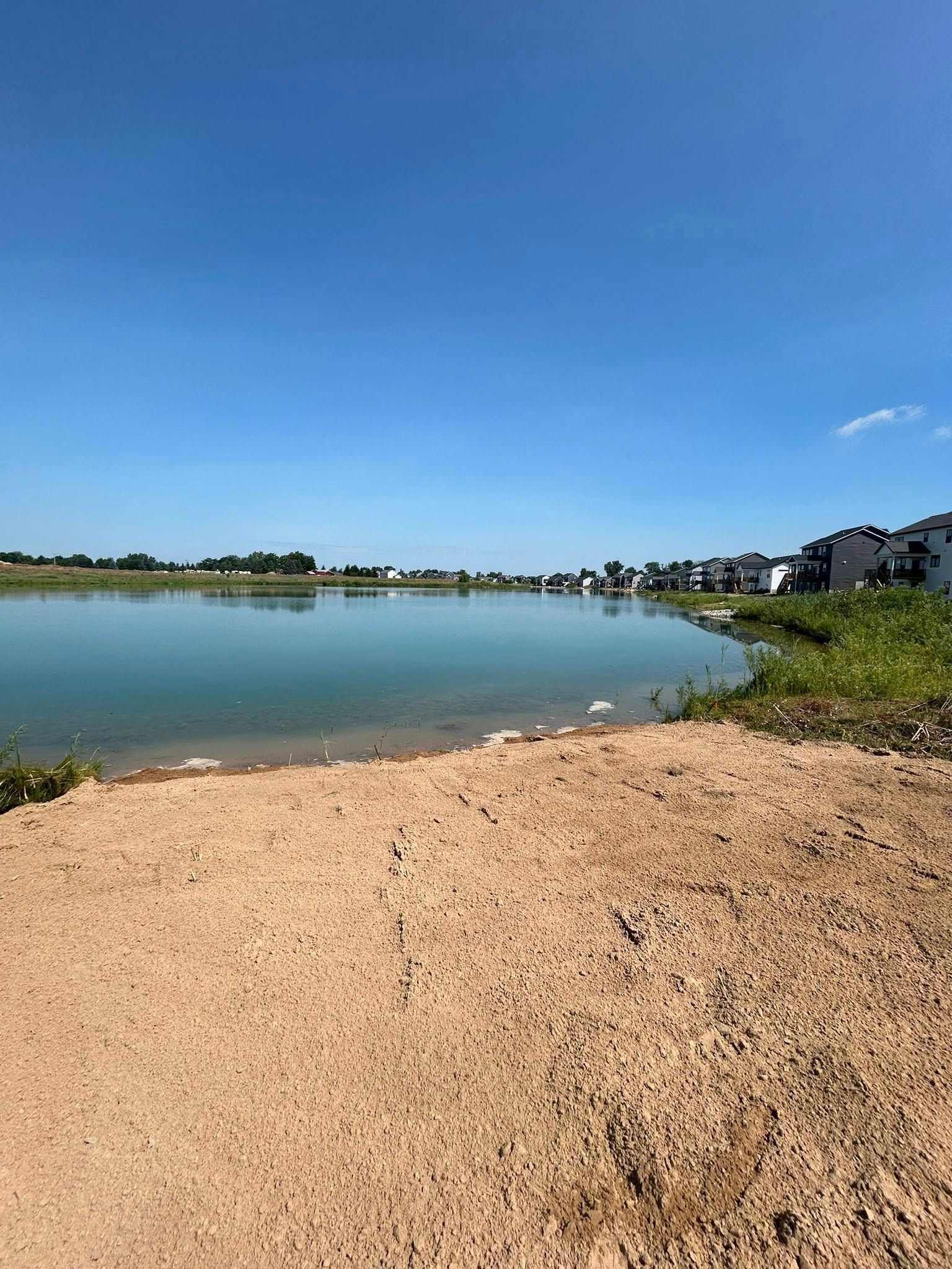 Sandy lakeshore under a clear blue sky, with calm water and houses along the opposite shore.