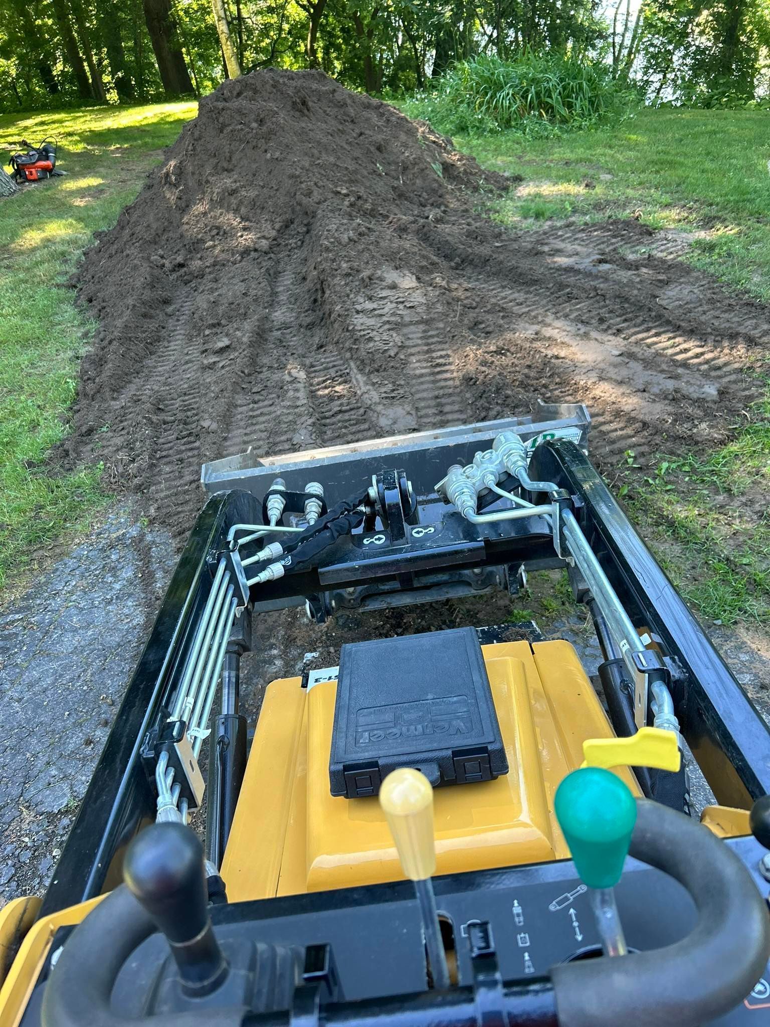 View from a small loader over a dirt pile in a grassy yard, with yellow controls in the foreground