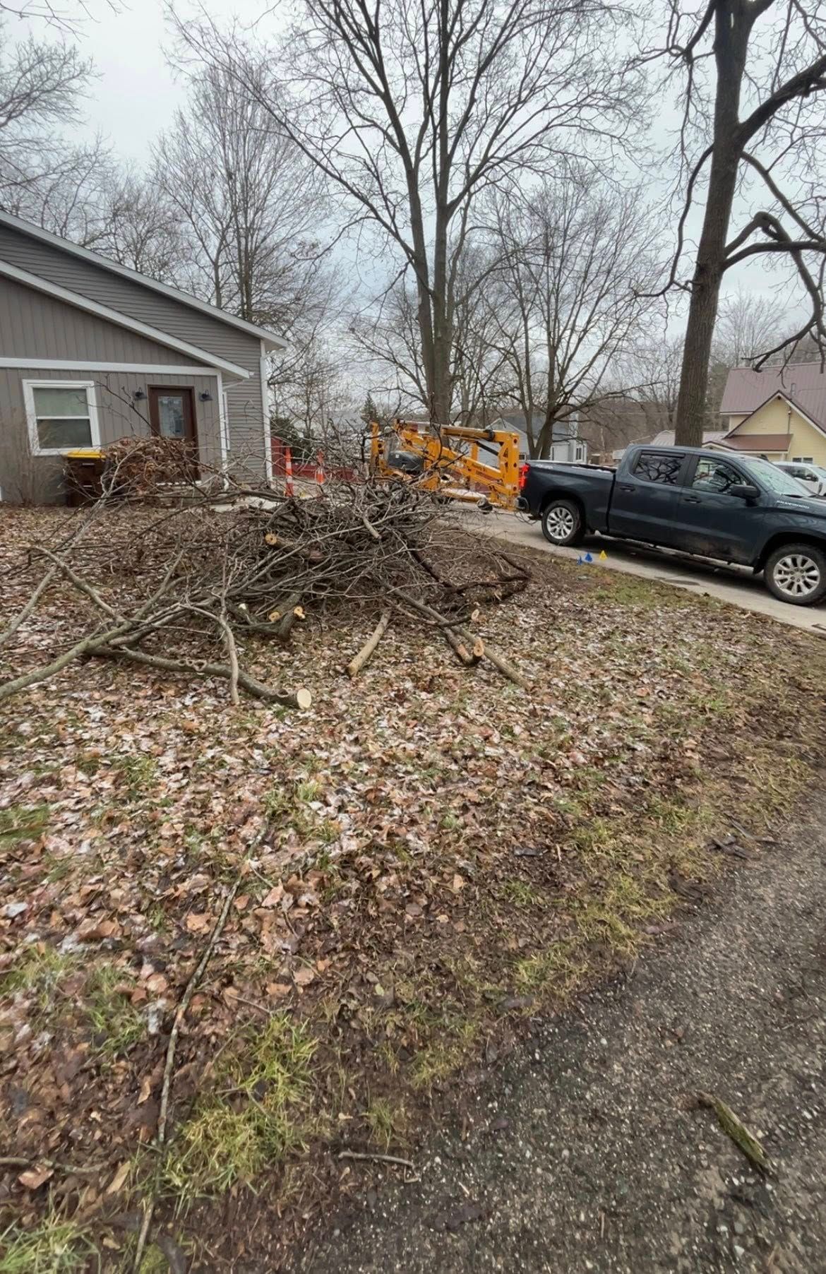Leaf-covered yard with a brush pile, parked black car, and orange utility truck beside a house.