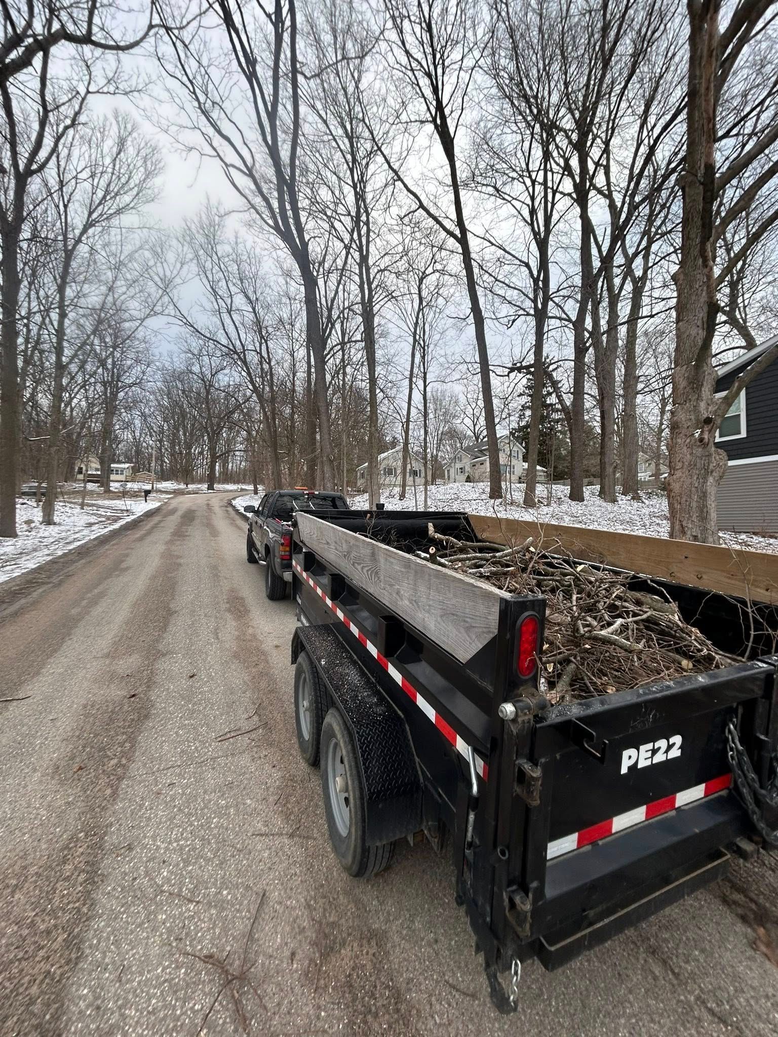 Black utility trailer loaded with gravel on a snowy gravel road lined with bare trees