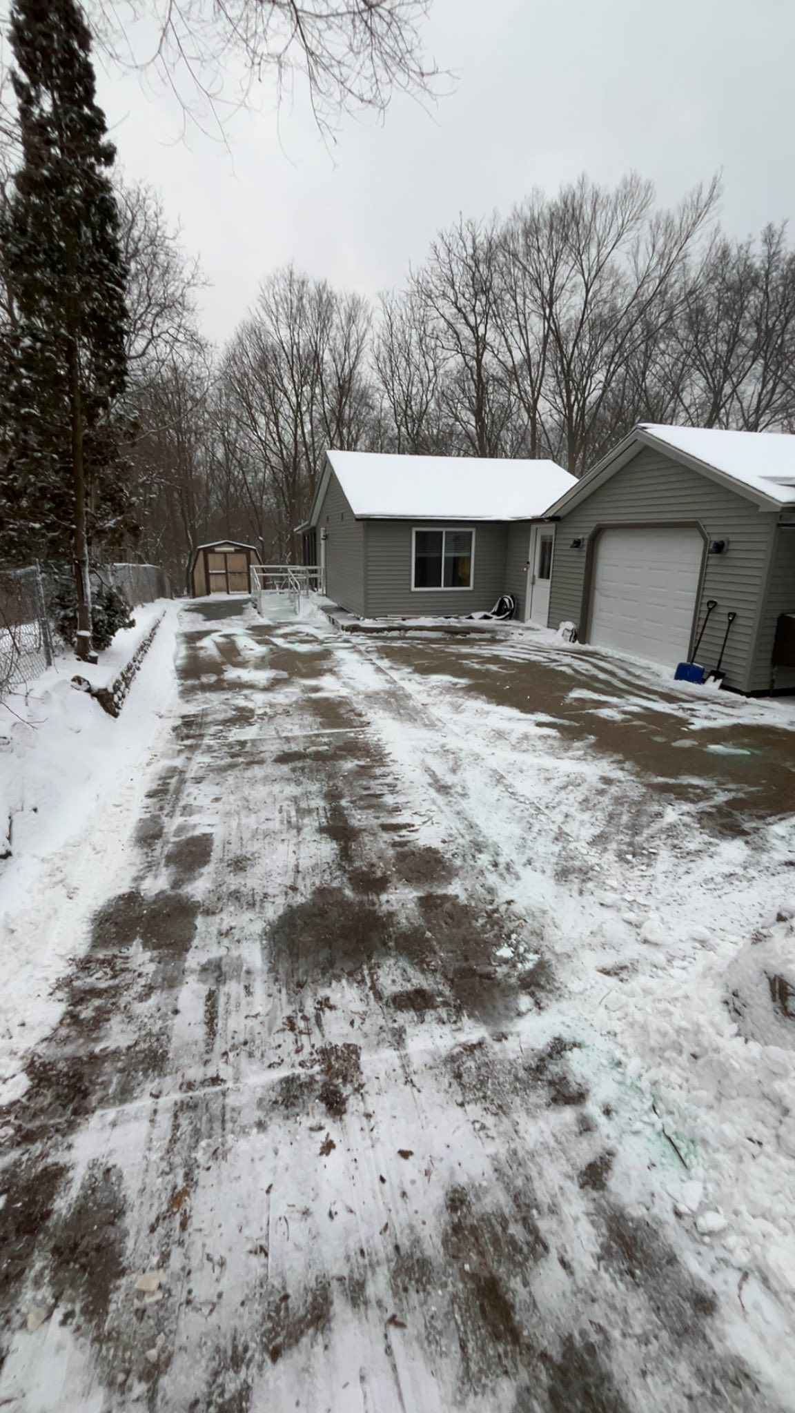 Snowy driveway beside a house and garage, with trees and light snow cover.