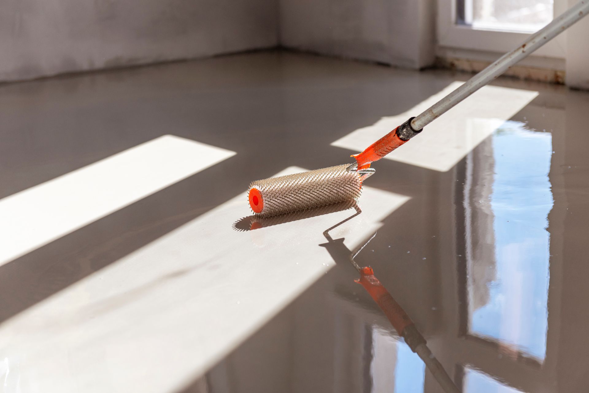 Une personne utilise un rouleau à peinture pour appliquer un revêtement gris clair sur un sol en béton à l'intérieur.