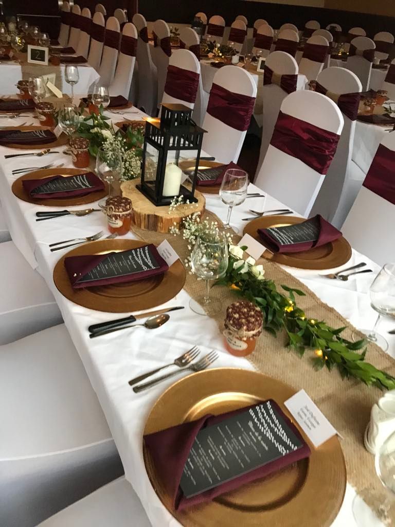 Une longue table est dressée pour une réception de mariage avec des assiettes dorées et des serviettes bordeaux.