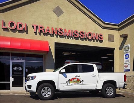 White pickup truck parked in front of Lodi Transmissions shop with red awning and sign, sunny day.