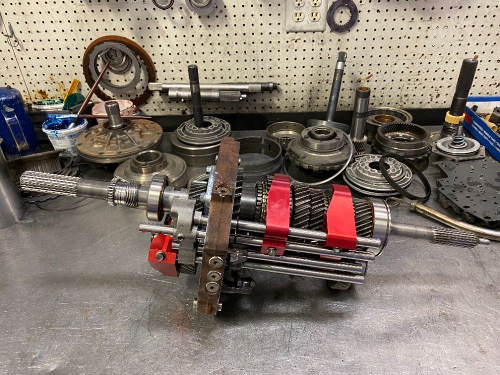 Disassembled transmission parts on a workbench, including gears, shafts, and housings. Shop environment, industrial setting.