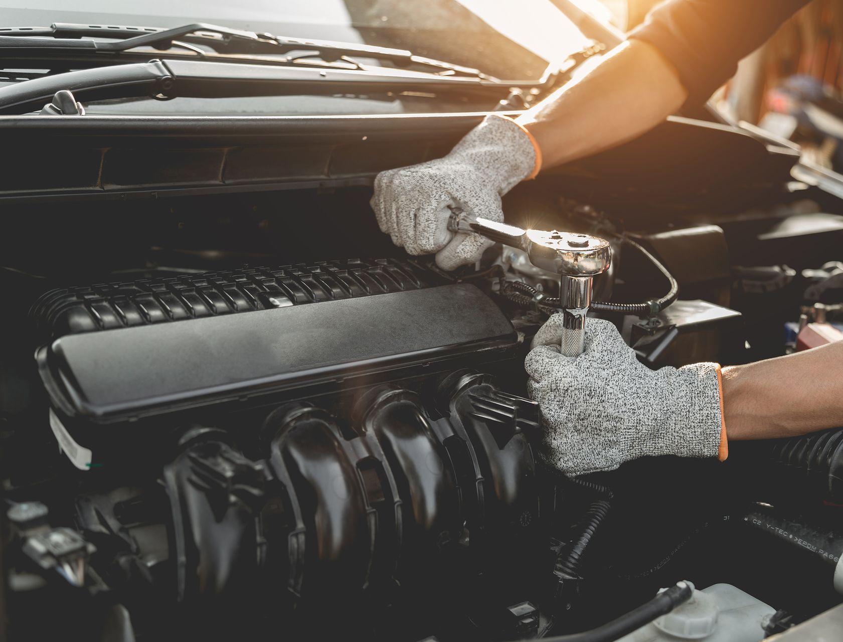 Mechanic using a wrench on a car engine, wearing work gloves. Bright sunlight shines on the engine bay.