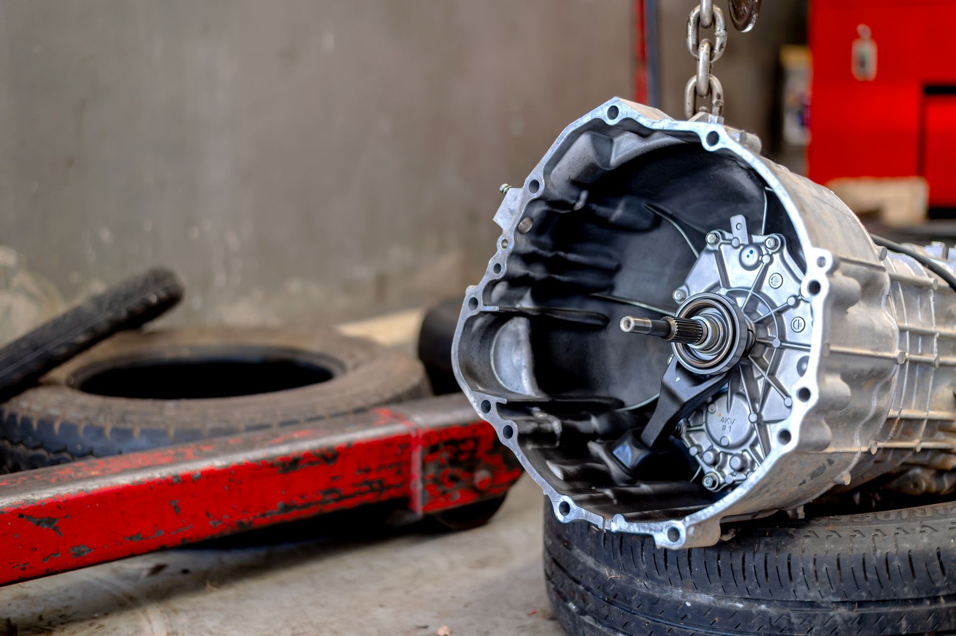 Open car transmission hanging, ready for repair, with tires and a red lift in the background.
