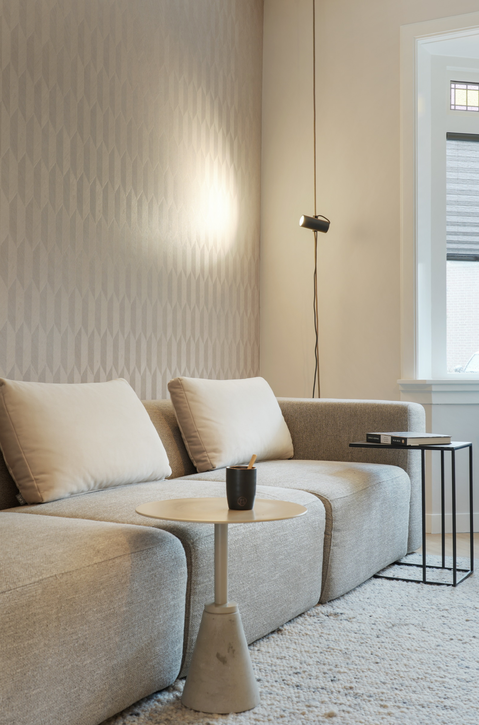 Gezellige woonkamer met grijze bank met structuur, behang met patroon, bijzettafels en hanglamp.