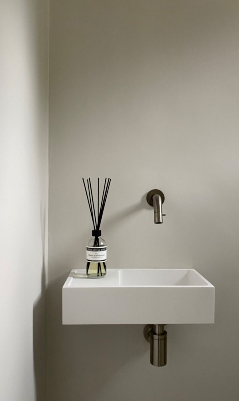 Wit Corian fonteintje met chromen kraan en afvoerbuis tegen een beige muur.