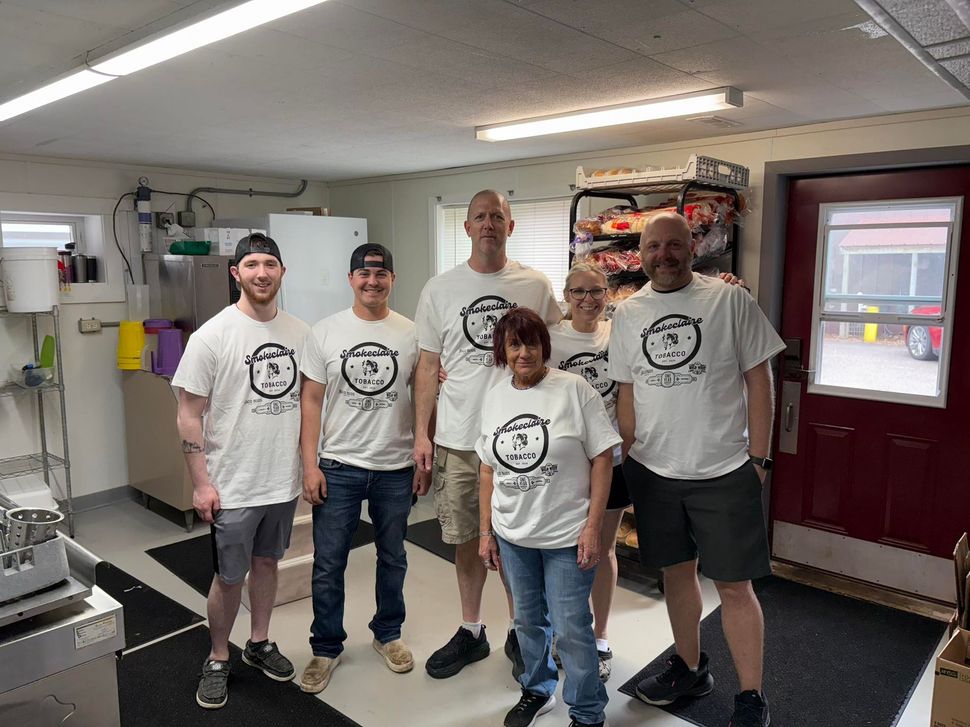 Six people in matching white team T-shirts pose for a photo inside a bright, industrial kitchen space.