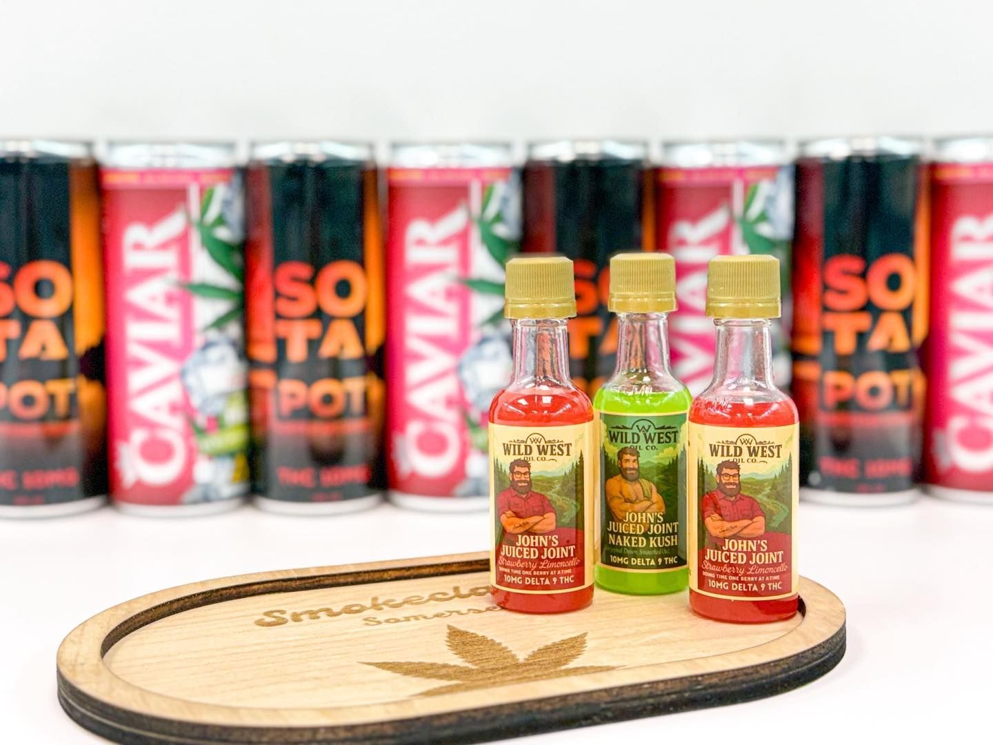 Three small liquid bottles stand on a wooden tray in front of a row of colorful canned beverages.