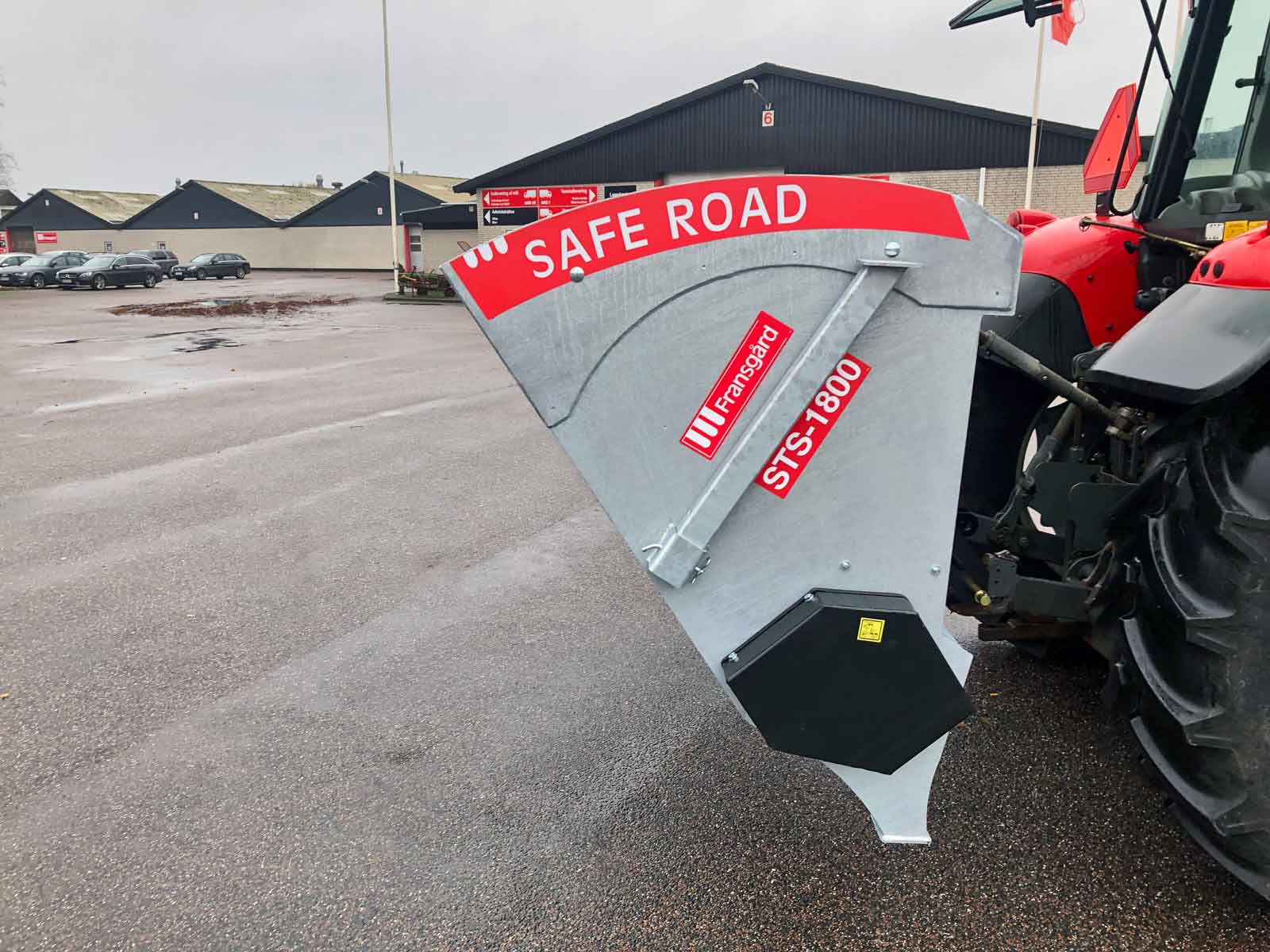 En rød traktor med en tallerkenspreder påmonteret på en parkeringsplads ved fabriksbygning.