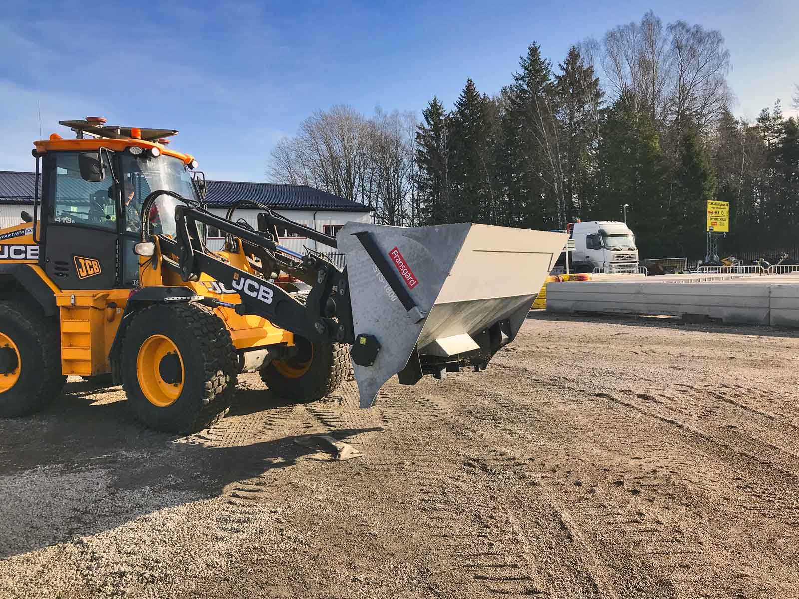 En gul og sort traktor med spreder monteret i grab kører på en grusplads.