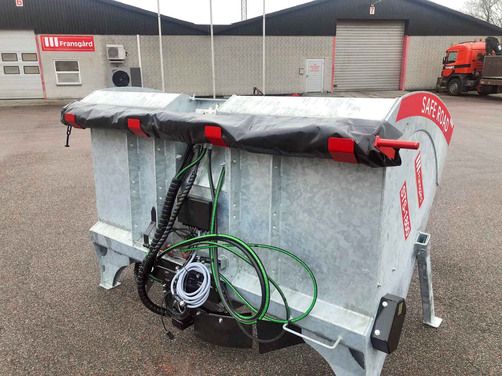 En tallerkenspreder med afmonteret pressenning er parkeret foran en bygning.