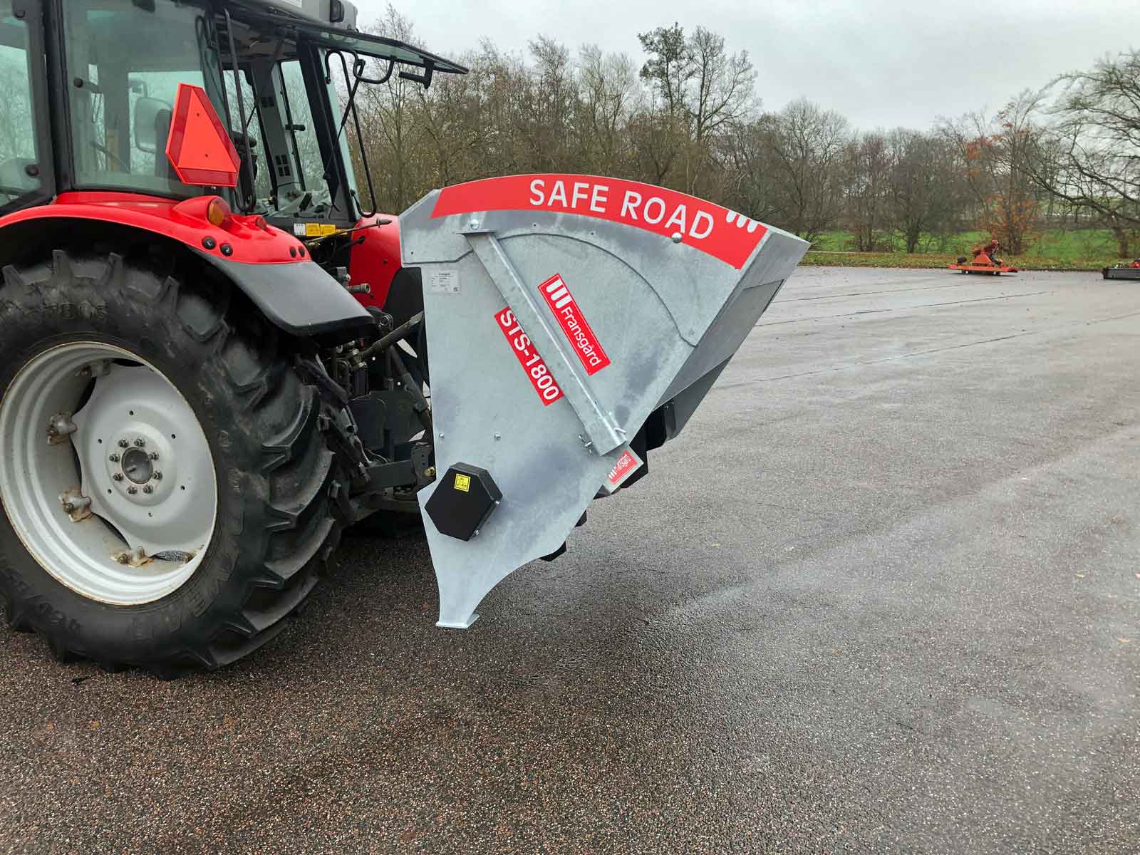 En rød traktor med en solid materialespreder fastgjort bagpå holder på en parkeringsplads.