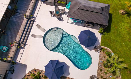 Overhead view of a backyard pool with blue water, patio umbrellas, lounge chairs, and a small gazebo