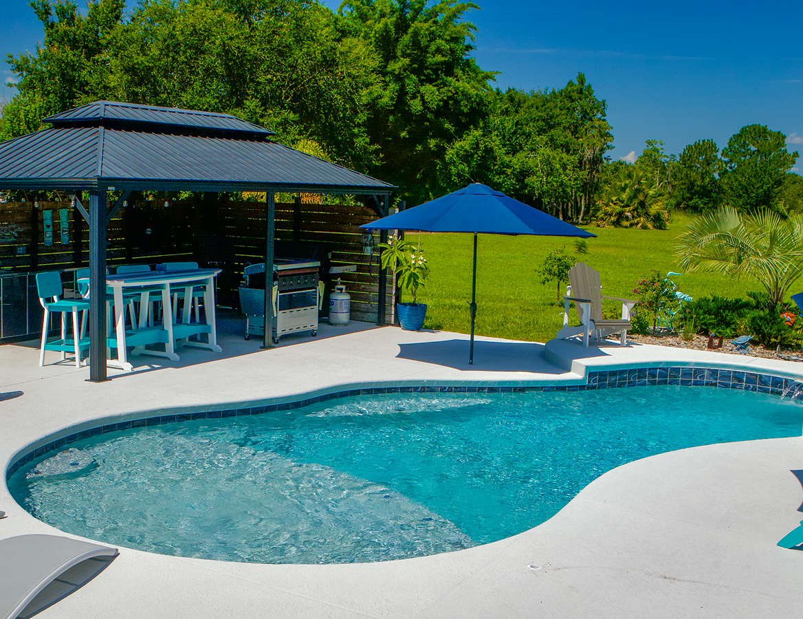 Backyard pool with blue umbrella, lounge chairs, and covered patio beside trees and lawn