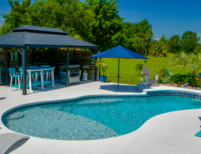 Backyard pool with blue umbrella, lounge chairs, and covered patio beside trees and lawn