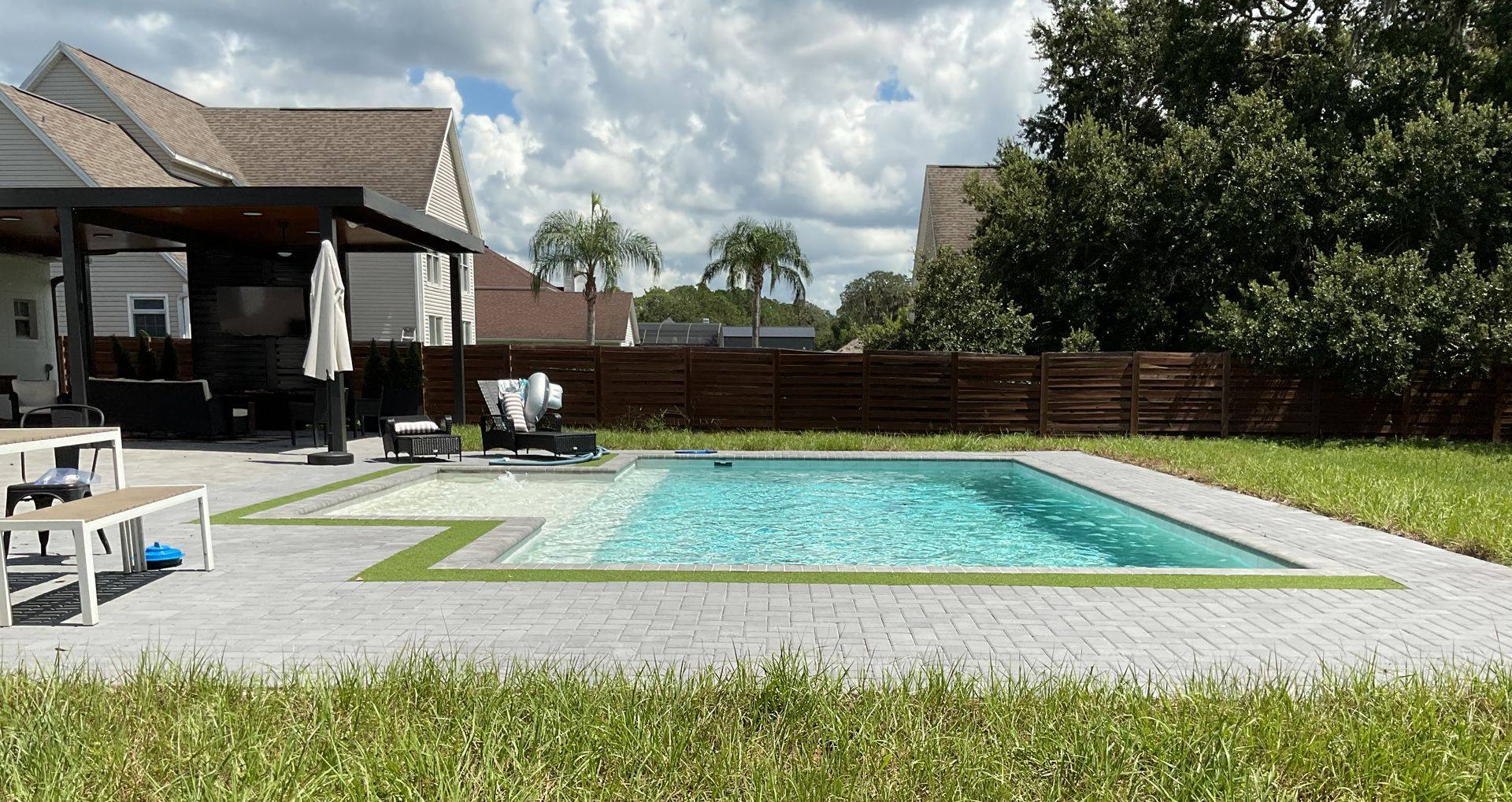 Backyard swimming pool beside a covered patio, with lounge chairs and a hedge fence.