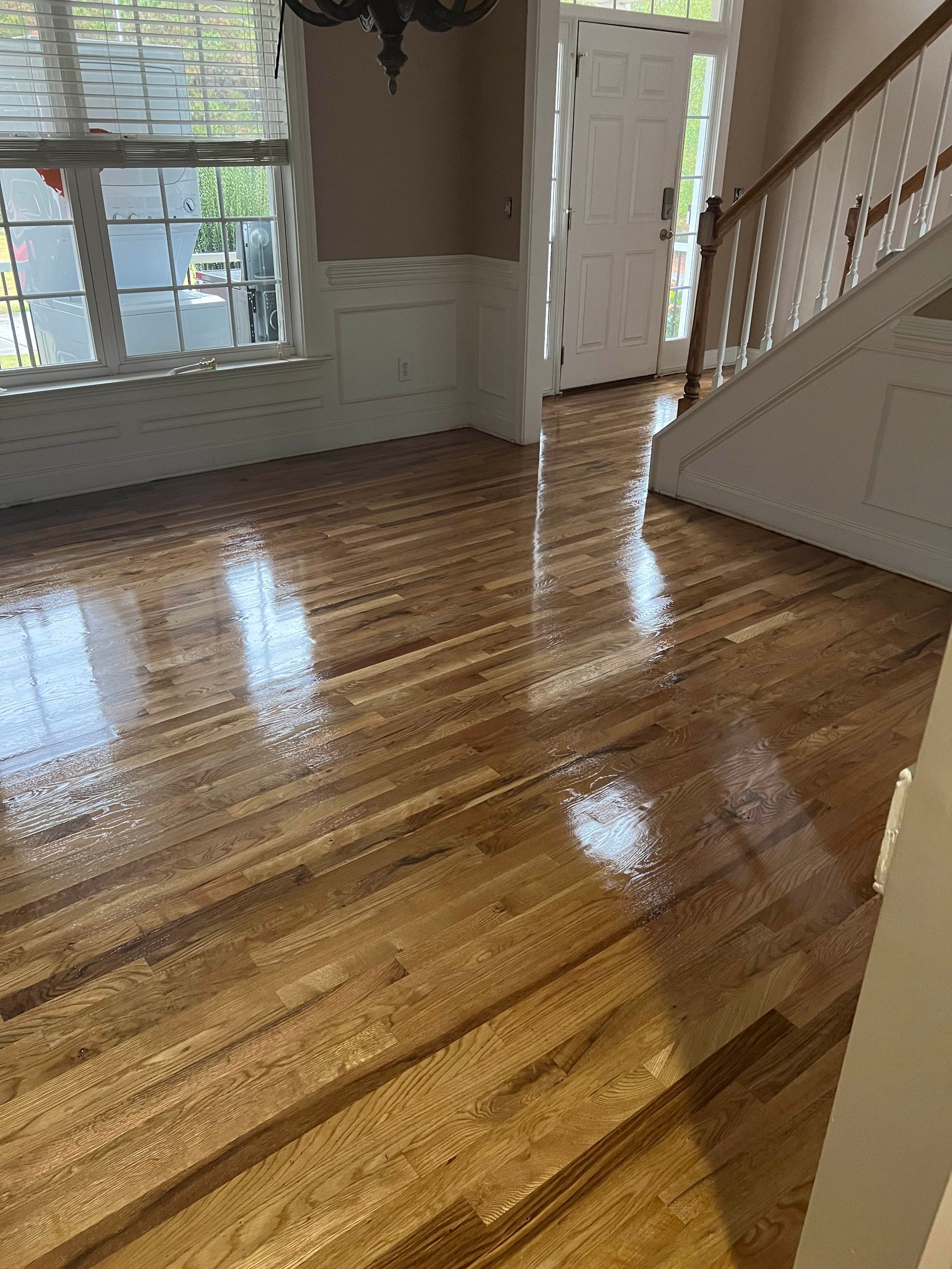 Empty sunlit room with glossy hardwood floors, staircase, and windows with white trim.