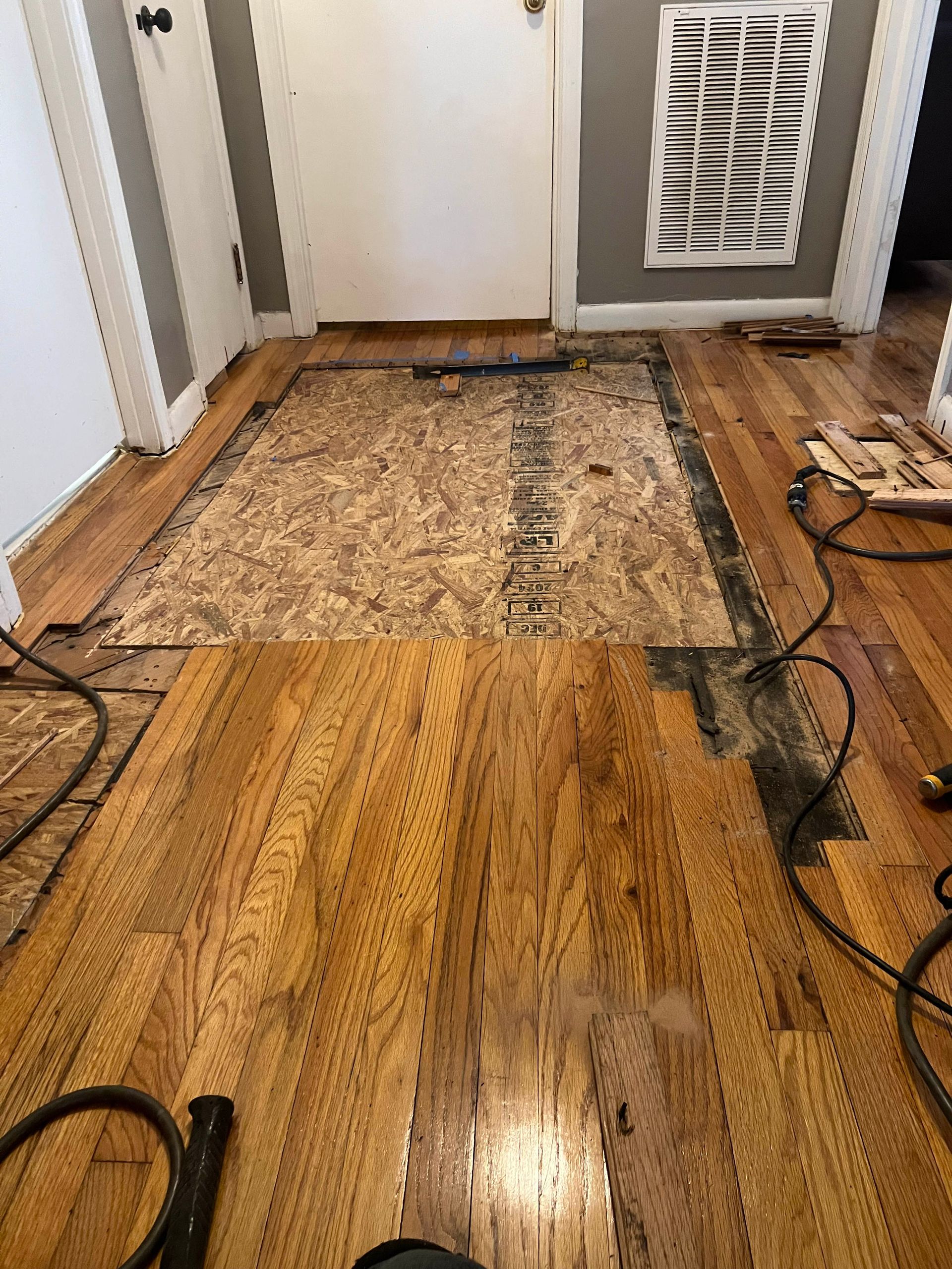 Damaged hardwood floor with dark stains and a worn rug near a doorway, with cords on the right.