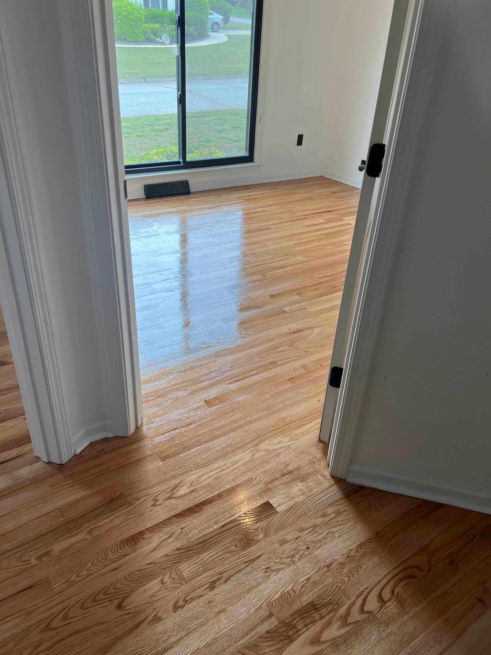 Empty hardwood-floored room with white walls and a large window overlooking greenery.