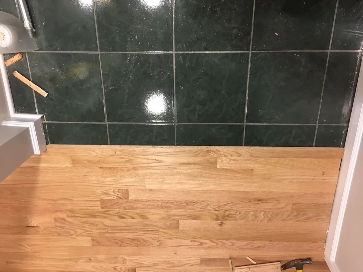 Wooden floor under construction beside dark tiled wall, with tools at the edge.