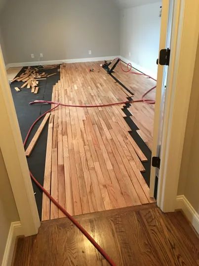 Room with hardwood floor under renovation, showing exposed boards, tools, and red hoses.