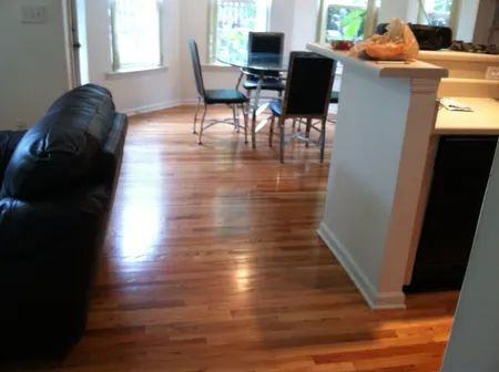 Bright living-dining room with hardwood floors, black sofa, glass table, chairs, and open kitchen counter.