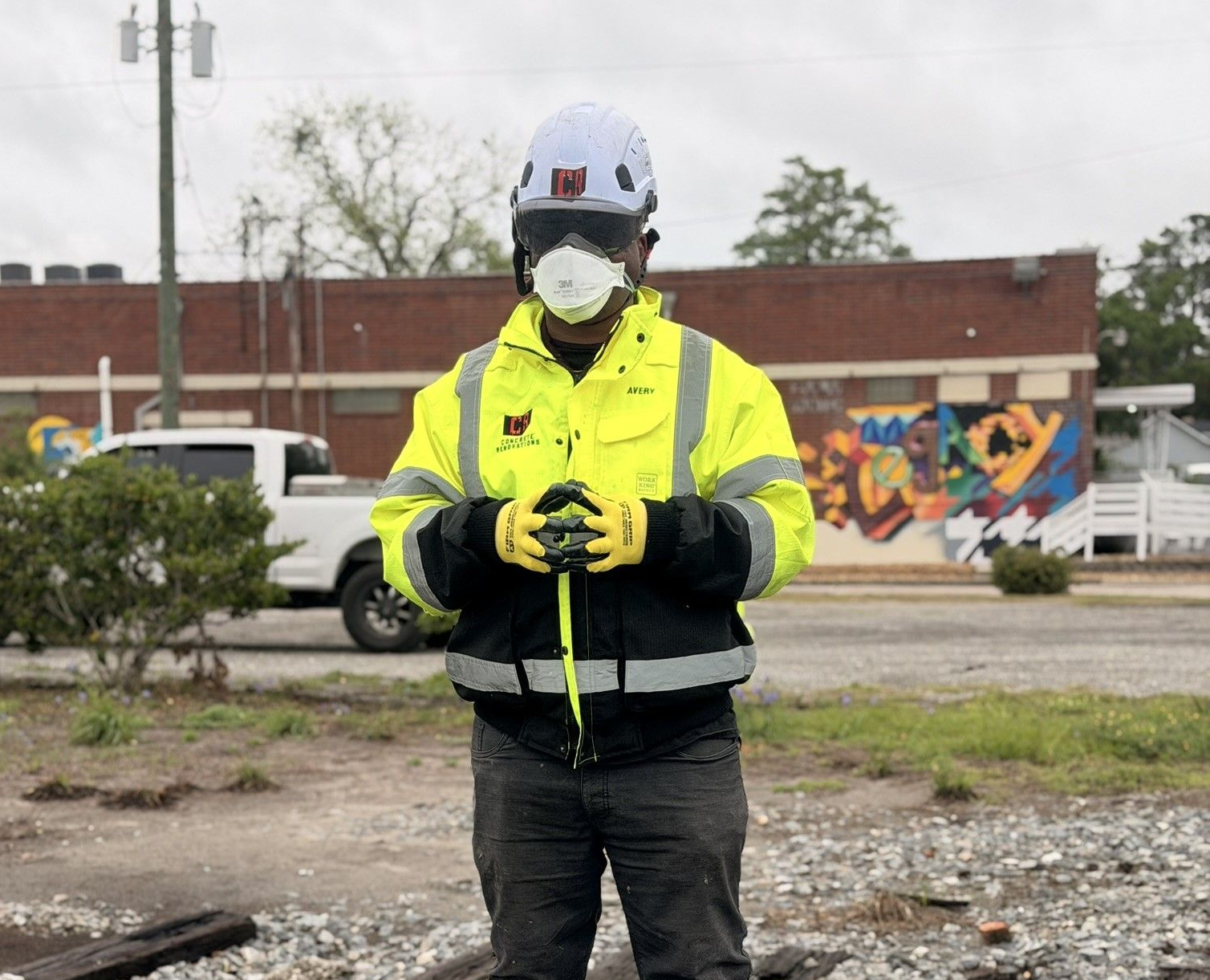 Worker in yellow safety jacket and helmet standing outdoors in front of a mural, holding a device.