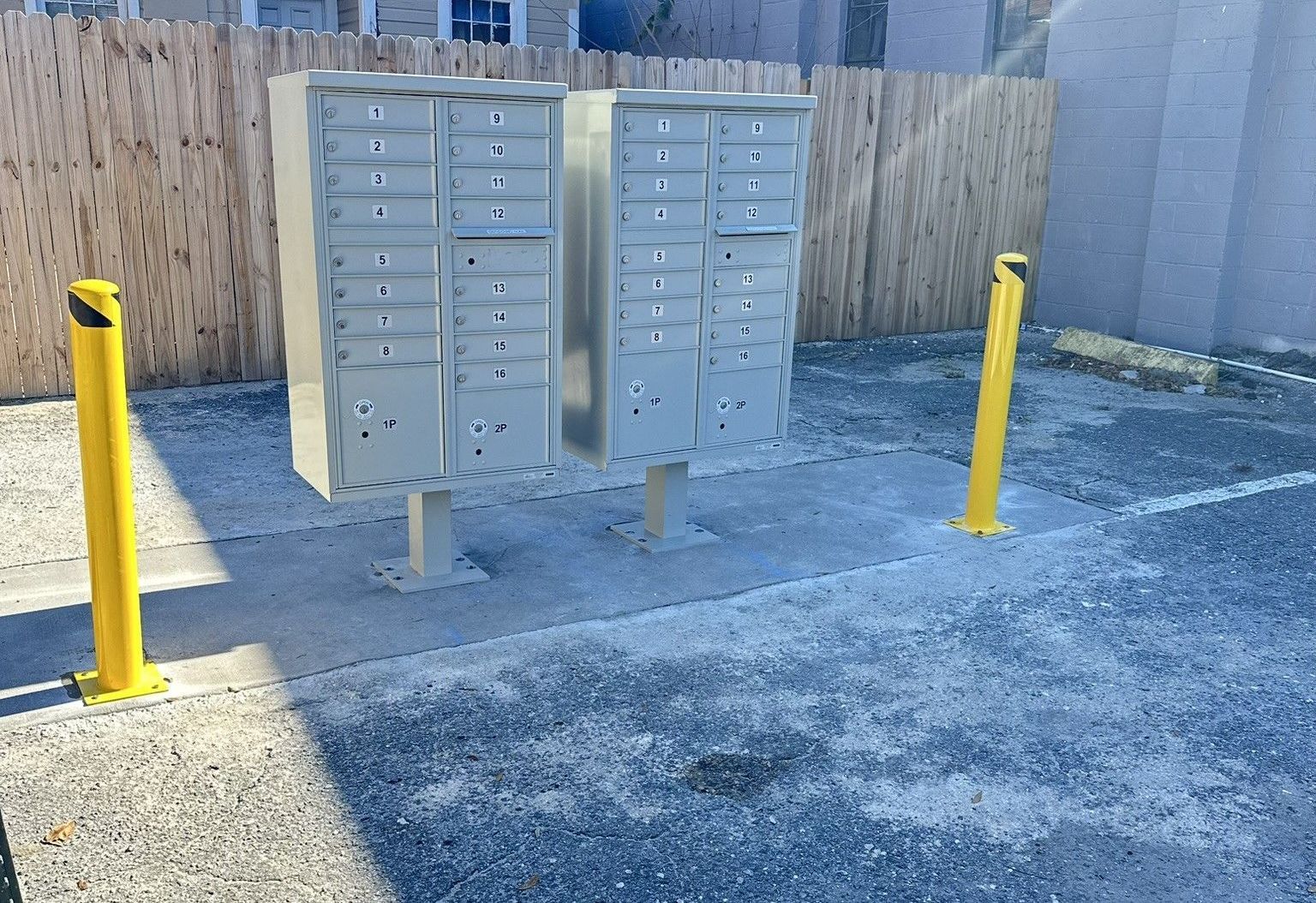 A row of mailboxes sitting on a concrete pad in a parking lot next to a wooden fence