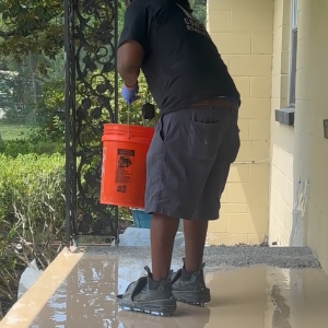 A man is holding an orange bucket and broadcasting vinyl flakes onto concrete porch