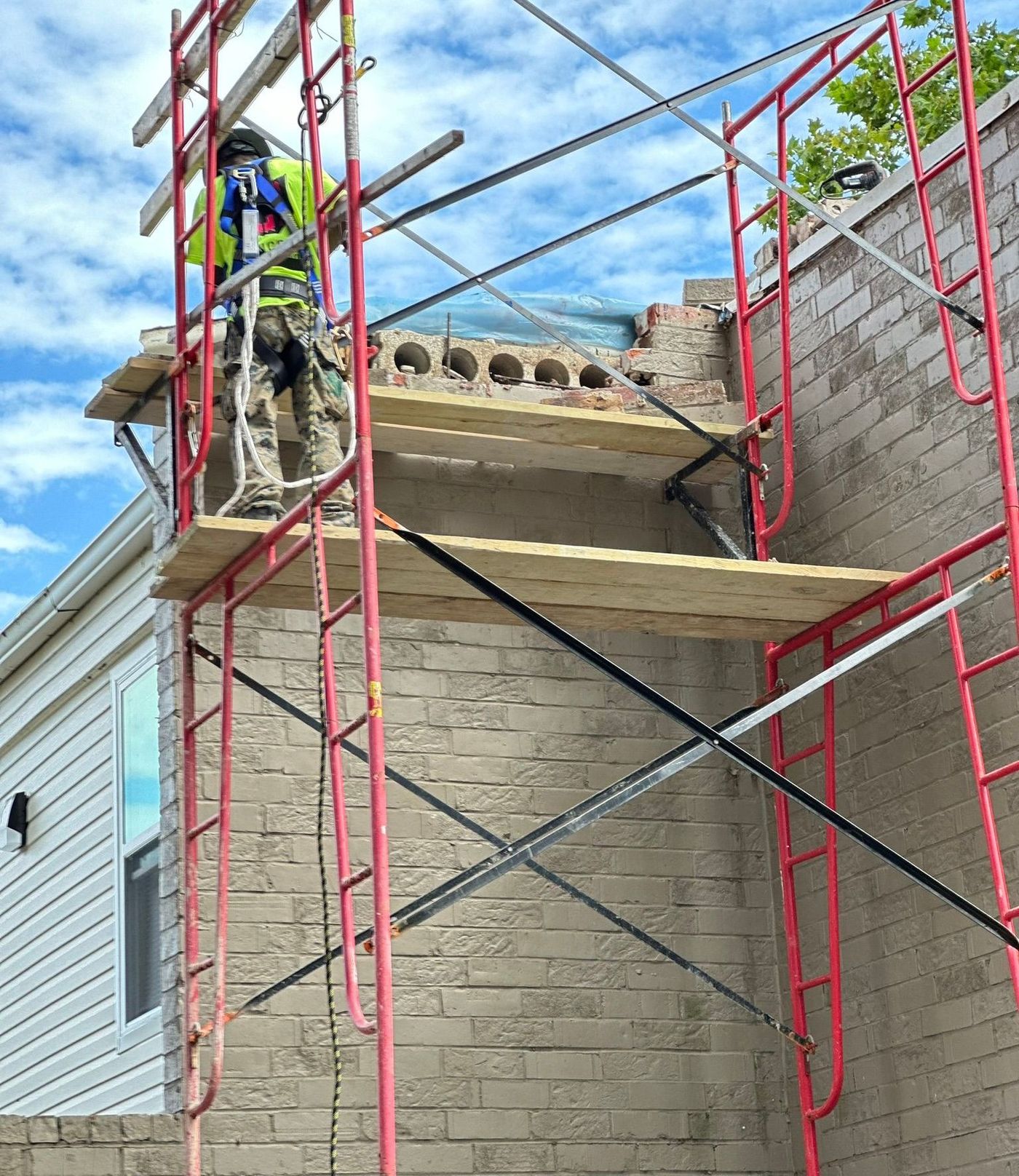 Worker on red scaffolding repairing a brick building roofline under a blue sky.