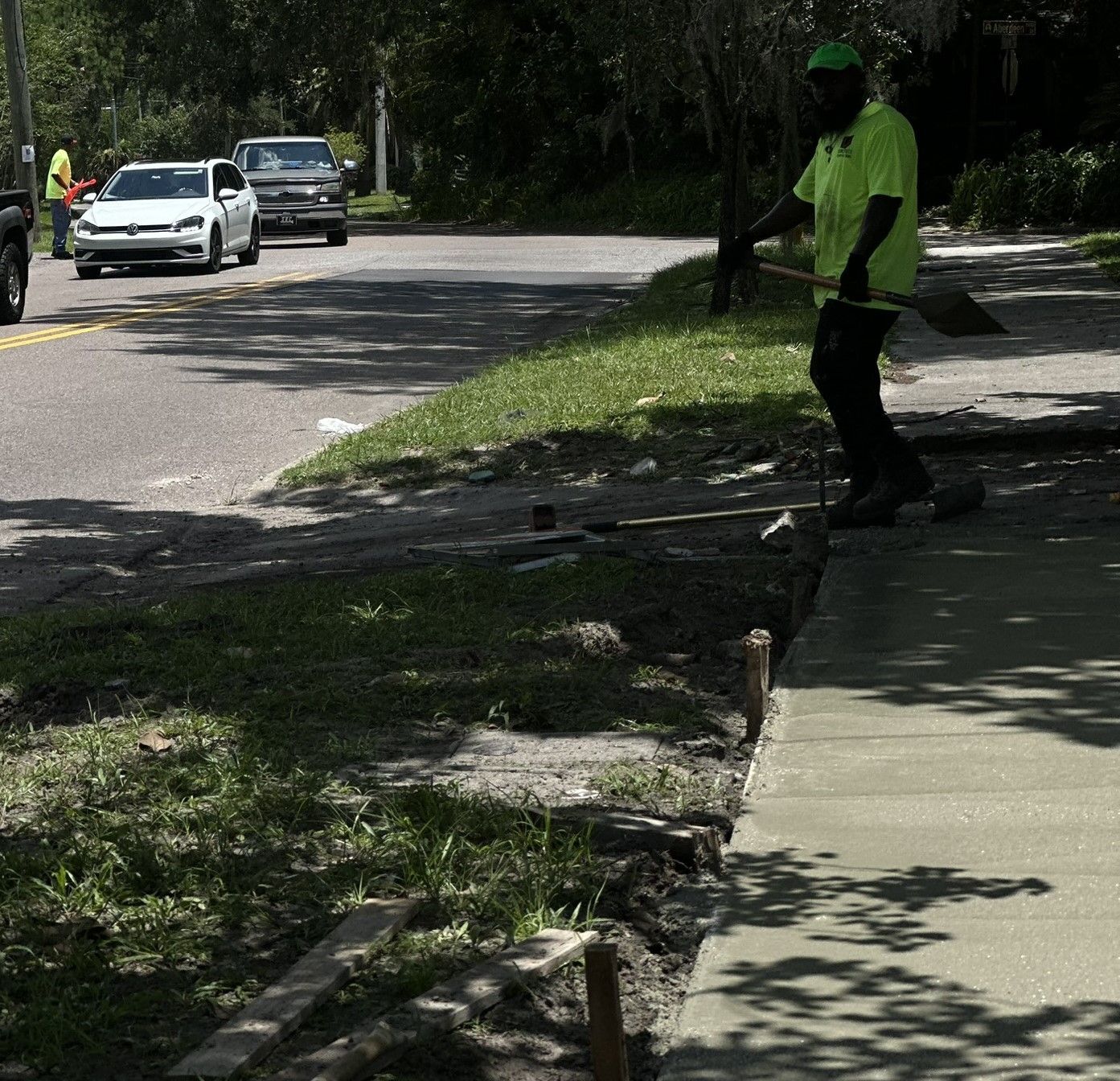 A man in a yellow shirt is standing on a concrete sidewalk holding a shovel