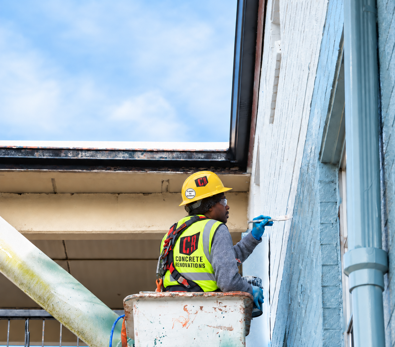 Worker in yellow hard hat and vest painting an exterior wall from a lift