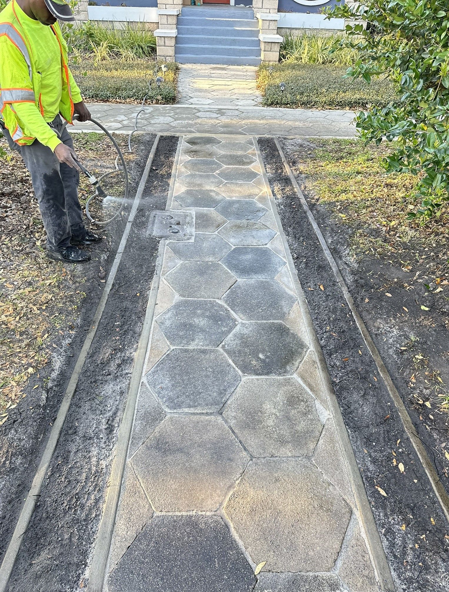 Worker pressure-washing a stone walkway bordered by shrubs and steps ahead