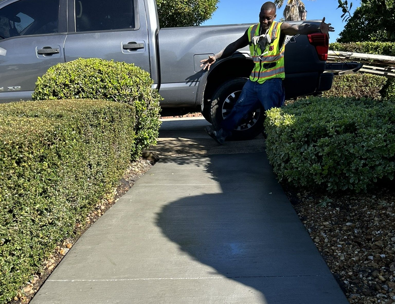 Worker in a safety vest standing beside a parked pickup truck on a sidewalk, next to a new concrete walkway, near trimmed hedges.