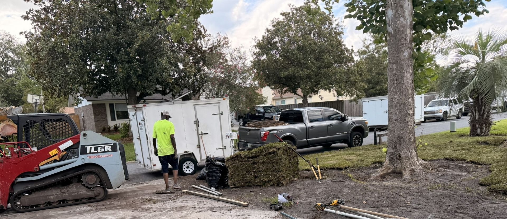 Worker and skid-steer loader clearing brush beside parked vehicles in a wooded lot