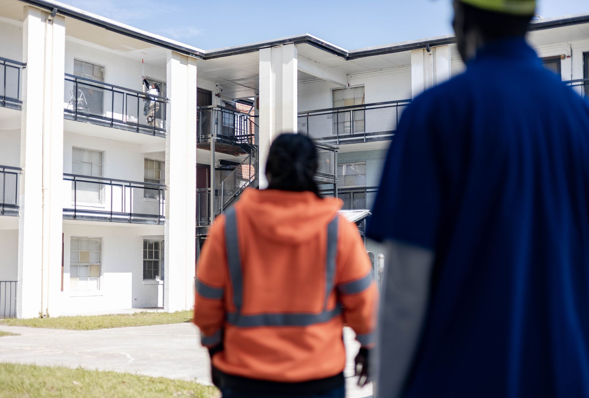 Avery McKnight in a safety vest walking towards a Concrete Renovations employee painting awhite apartment building with black balconies.