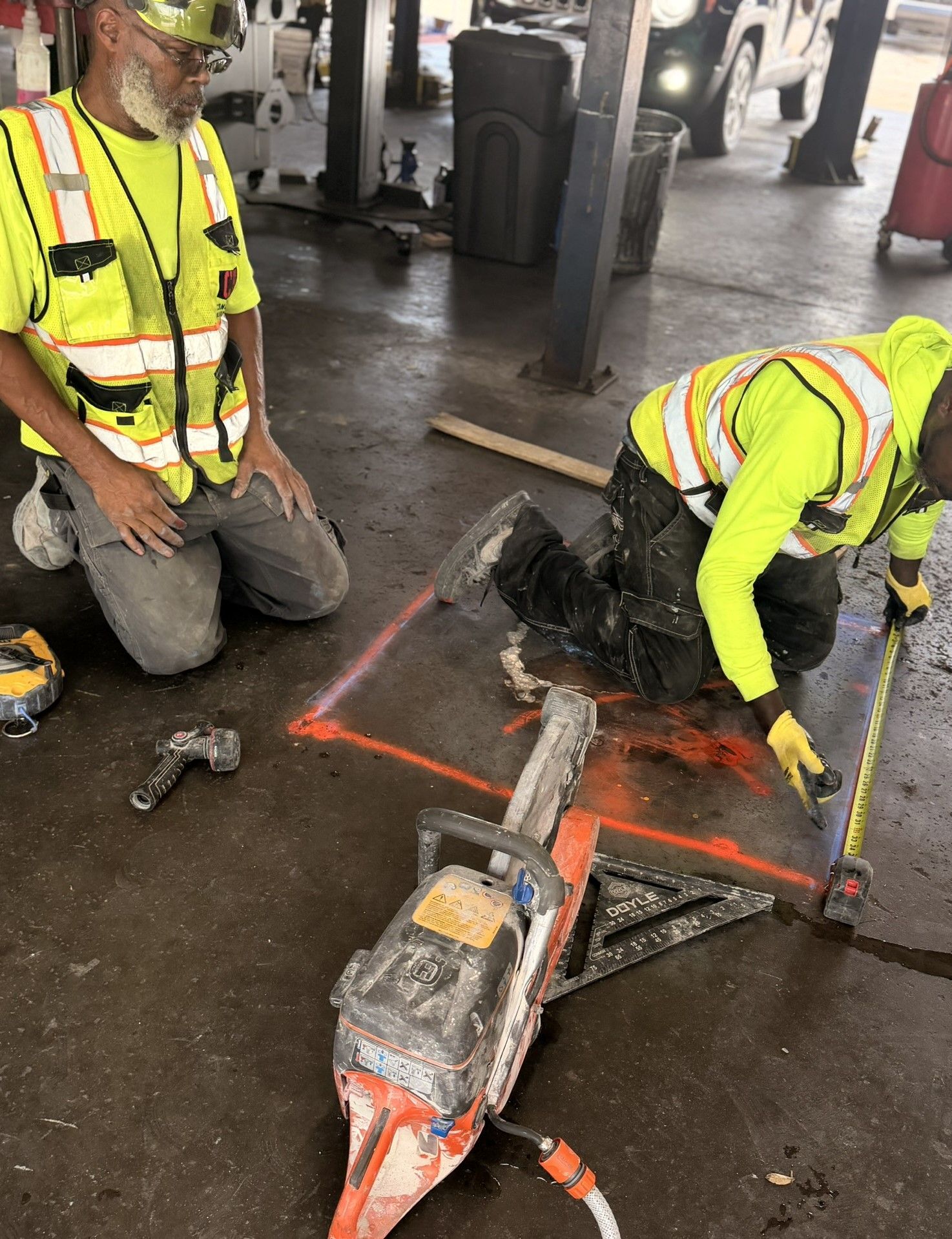 Two workers in high-vis gear cut pavement with a concrete saw under a covered work area.