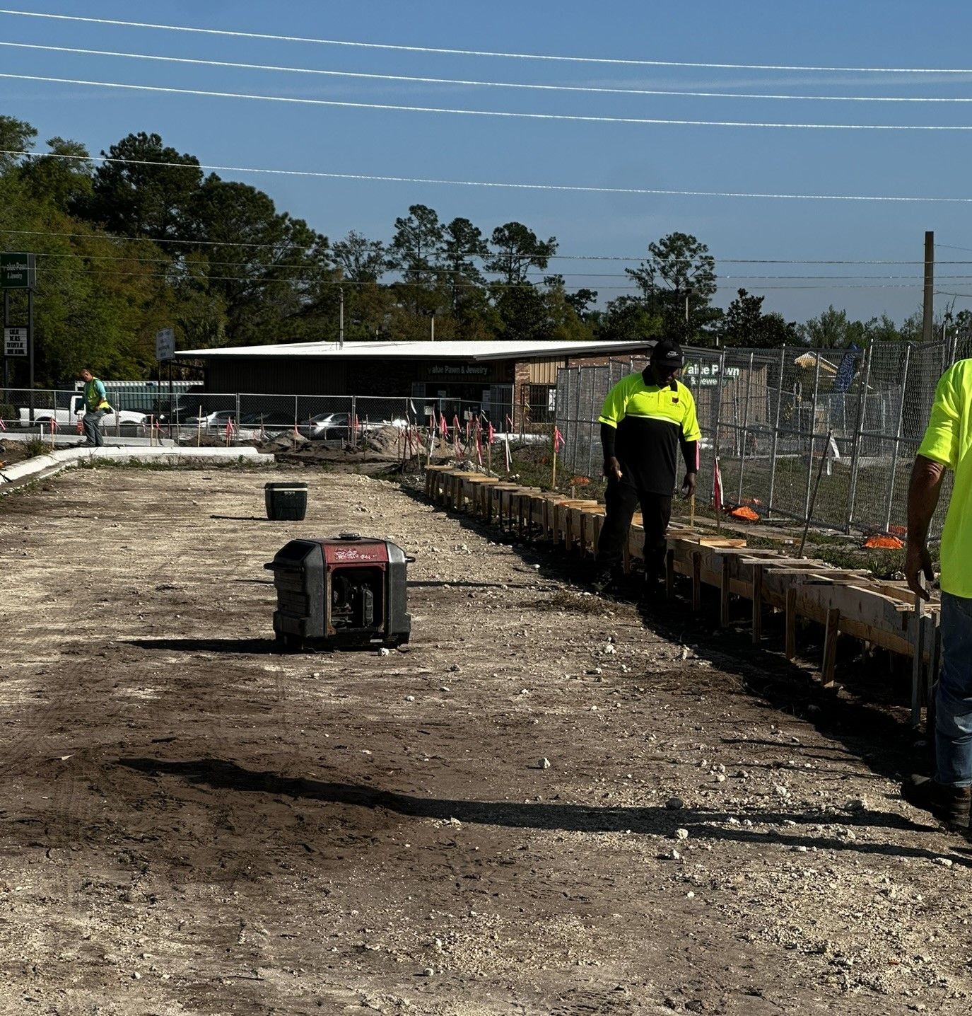 Construction workers in neon vests on a dirt worksite beside a fence under a clear blue sky.