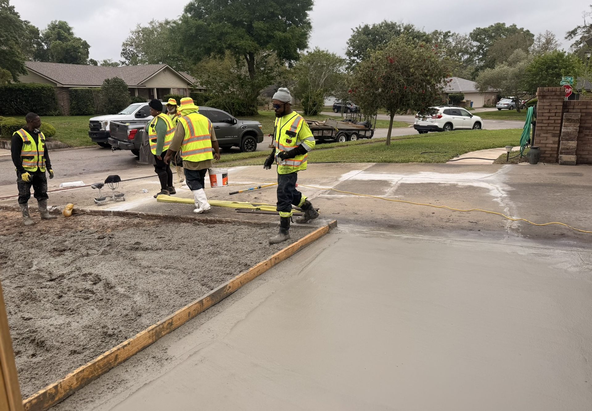 Workers in yellow safety gear pour fresh concrete at a suburban curbside construction site.