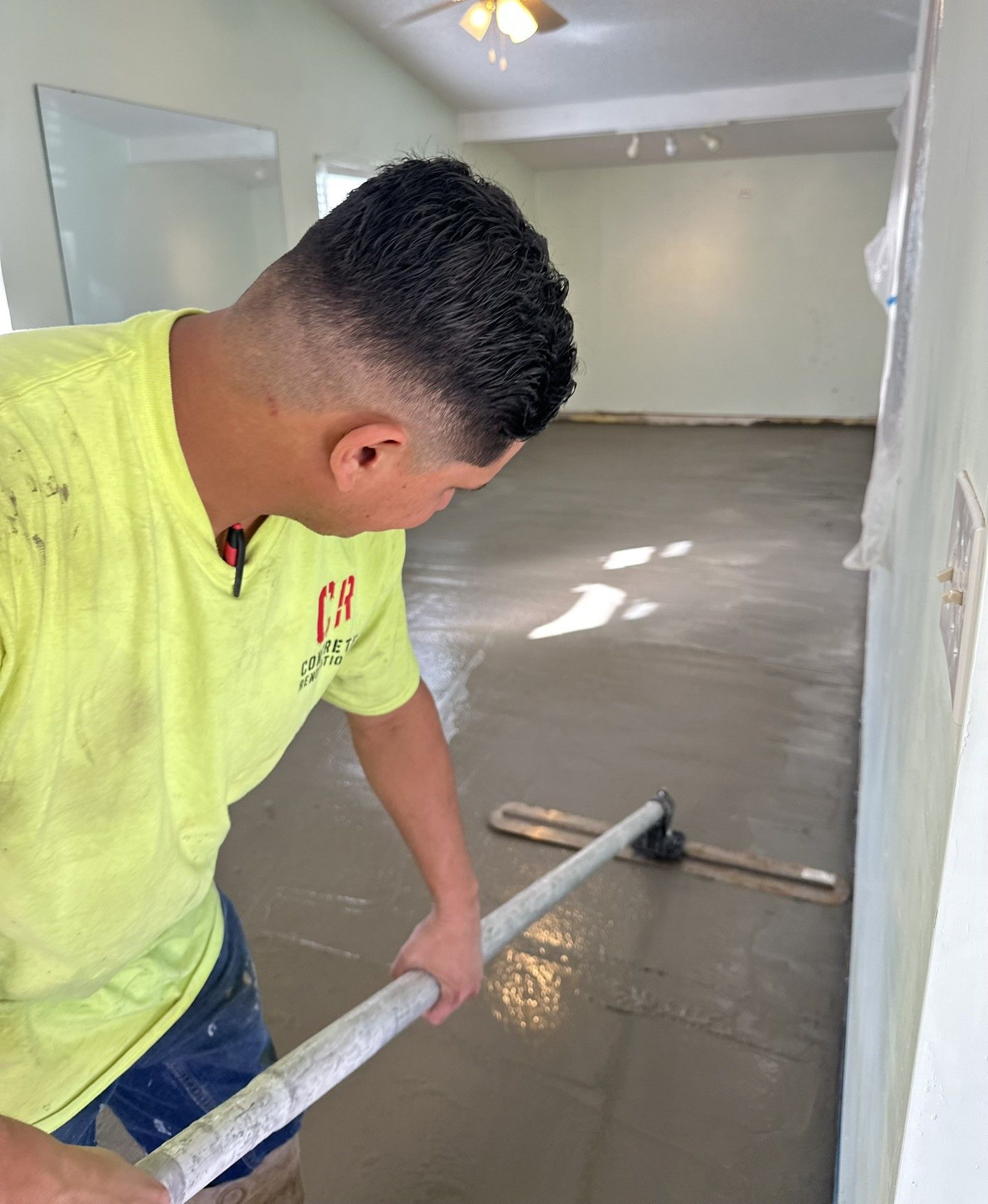 Man smoothing wet concrete floor with a long leveler in a room with light green walls.
