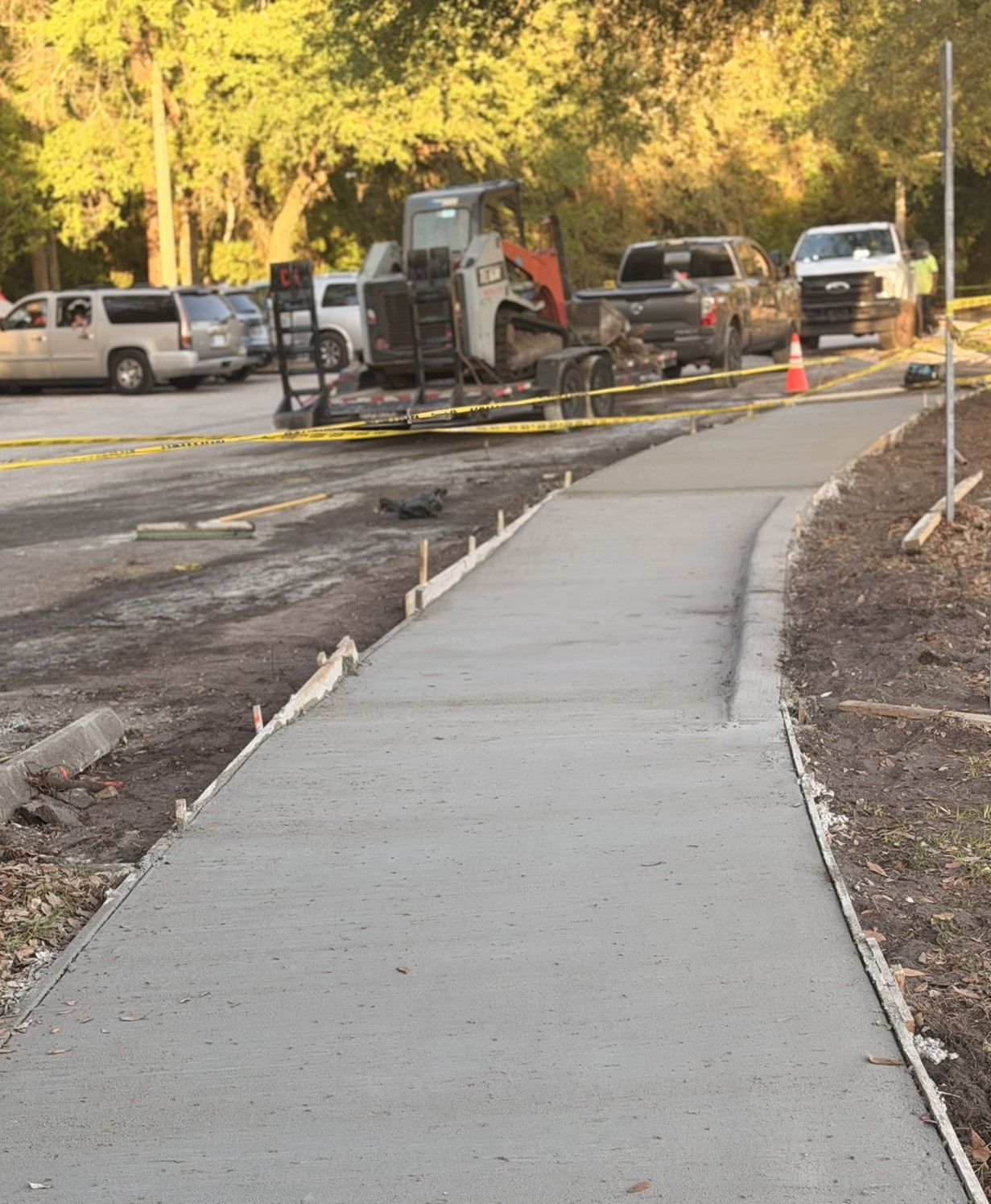 Curved sidewalk beside a construction area with vehicles, caution tape, and orange cones.