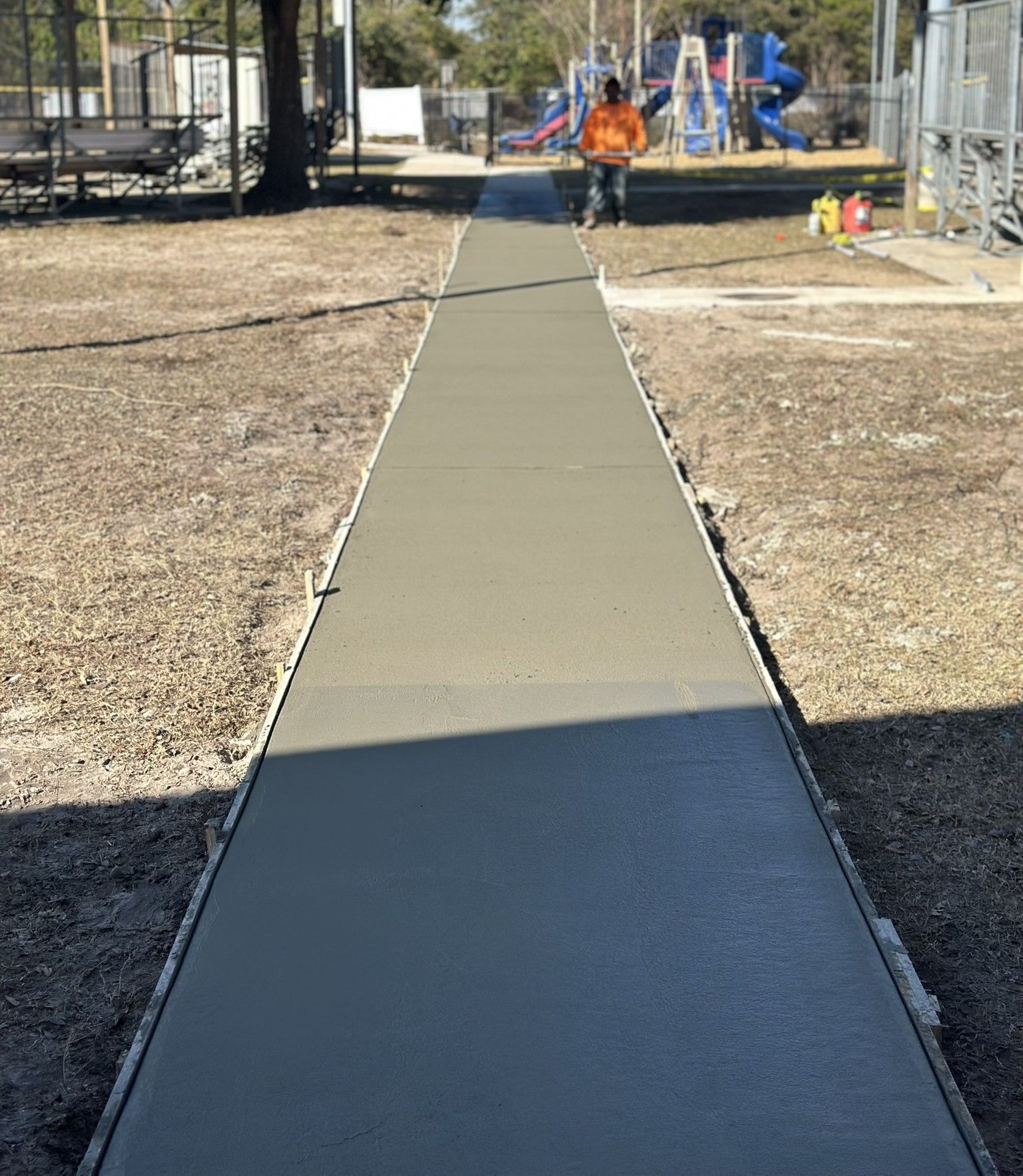 Fresh concrete walkway through a park, with a worker in orange near a playground.